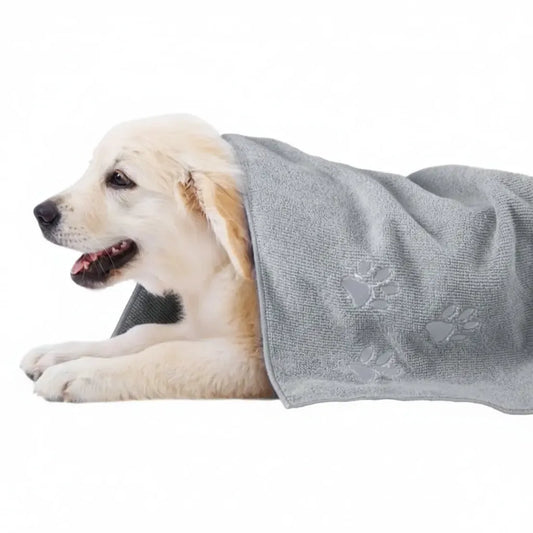 A light cream-colored puppy lying on a white background, wrapped in a light gray textured pet towel with embossed paw prints.