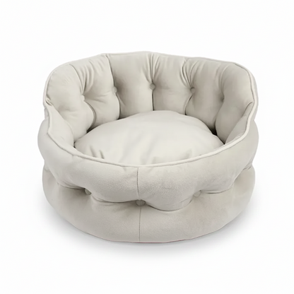 A round tufted pet bed in a light beige or cream faux-leather fabric, featuring a high bolstered wall and a matching cushion inside, shown on a plain white background.