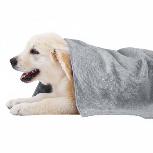 A light cream-colored puppy lying on a white background, wrapped in a light gray textured pet towel with embossed paw prints.